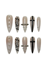 Set of long stiletto-shaped nude press-on nails with silver chrome studs, black gothic typography, and cross motifs. Features brutalist nail art with edgy glam detailing for alternative fashion styles.